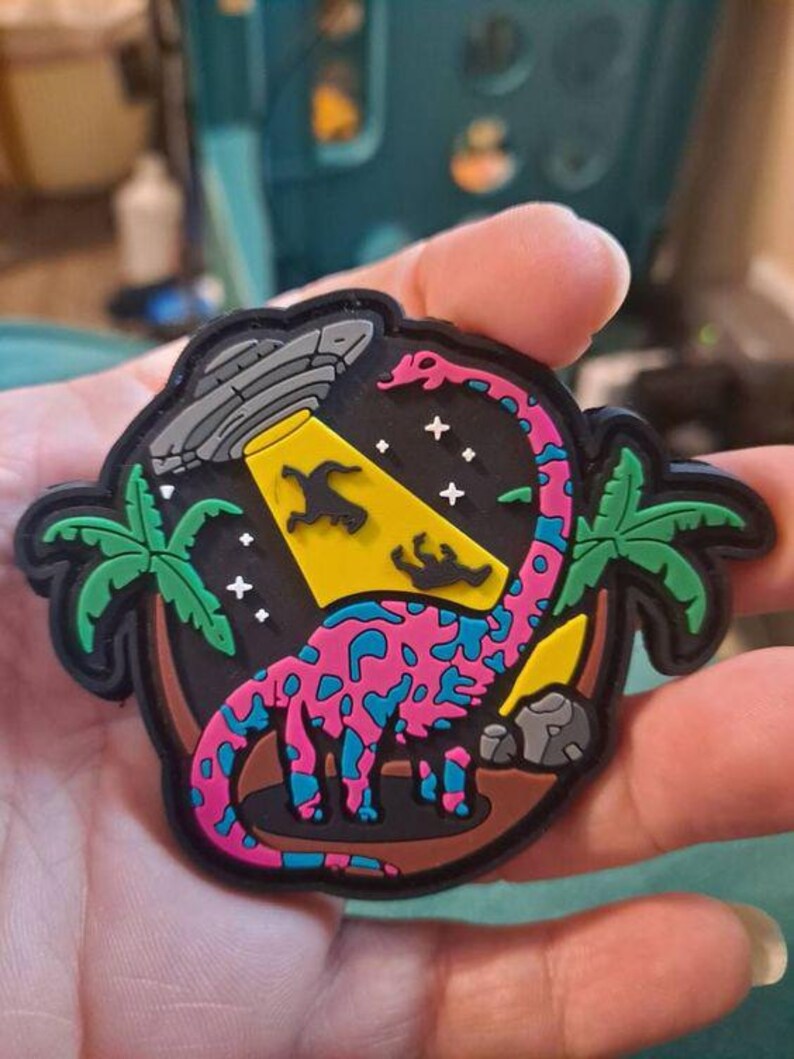 Może przedstawiać: Kolorowa naszywka przedstawiająca r&oacute;żowego i niebieskiego dinozaura porwanego przez UFO. Projekt zawiera palmy, gwiazdy i ż&oacute;łty reflektor. Naszywka ma czarną obw&oacute;dkę i brązowe tło.