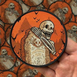 Może przedstawiać: Okrągła naszywka z czarną obw&oacute;dką przedstawiająca szkielet oparty o nagrobek z napisem "Memento Mori". Tło jest pomarańczowe z g&oacute;rami i nietoperzami.
