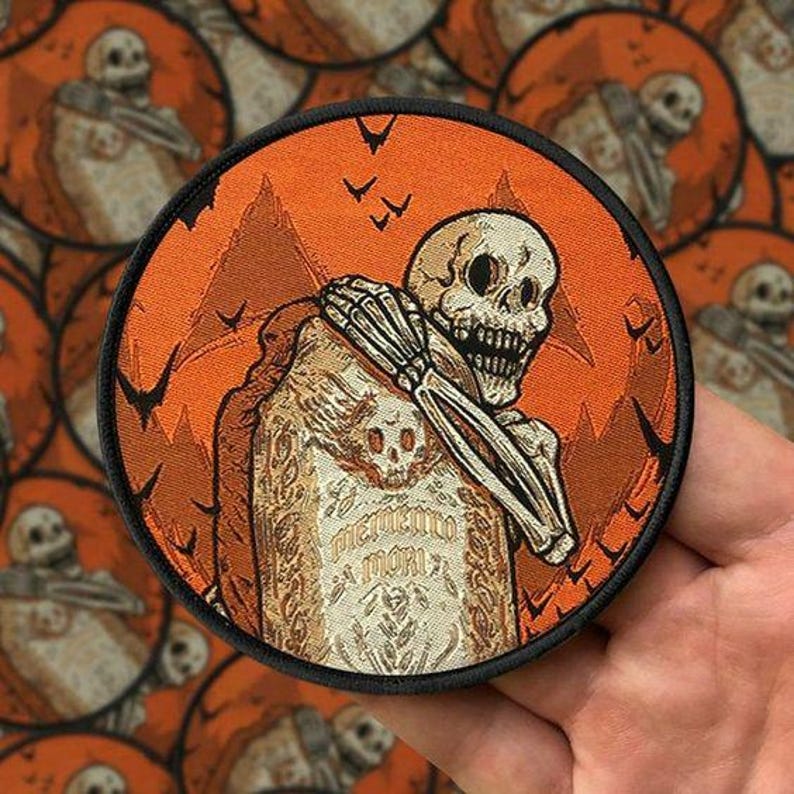 Może przedstawiać: Okrągła naszywka z czarną obw&oacute;dką przedstawiająca szkielet oparty o nagrobek z napisem "Memento Mori". Tło jest pomarańczowe z g&oacute;rami i nietoperzami.