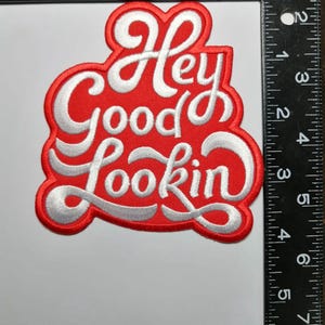 Może przedstawiać: Czerwona, haftowana naszywka z napisem "Hey Good Lookin" w białym skrypcie. Naszywka ma około 10 cm szerokości i 10 cm wysokości.