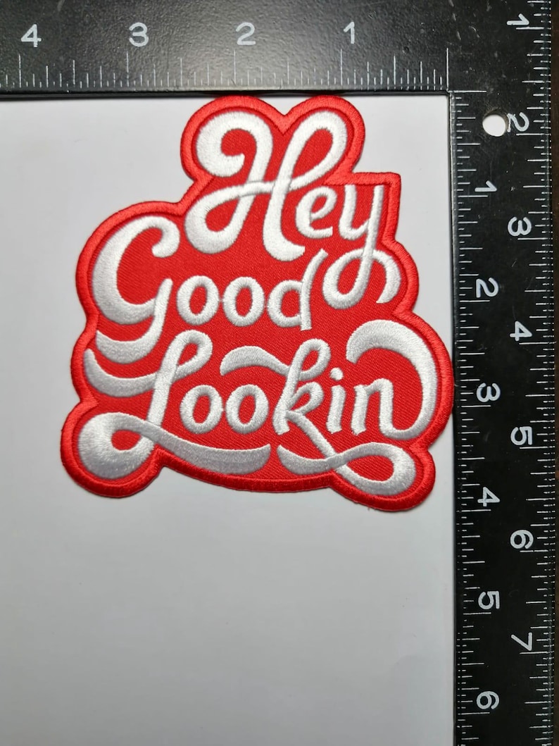 Może przedstawiać: Czerwona, haftowana naszywka z napisem "Hey Good Lookin" w białym skrypcie. Naszywka ma około 10 cm szerokości i 10 cm wysokości.