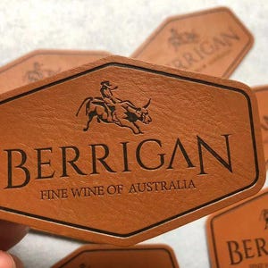 Może przedstawiać: Brązowa sk&oacute;rzana etykieta na wino z napisem "BERRIGAN" i tekstem "FINE WINE OF AUSTRALIA". Etykieta ma czarną obw&oacute;dkę i grafikę przedstawiającą osobę jadącą na byku. Etykieta ma sześciokątny kształt.