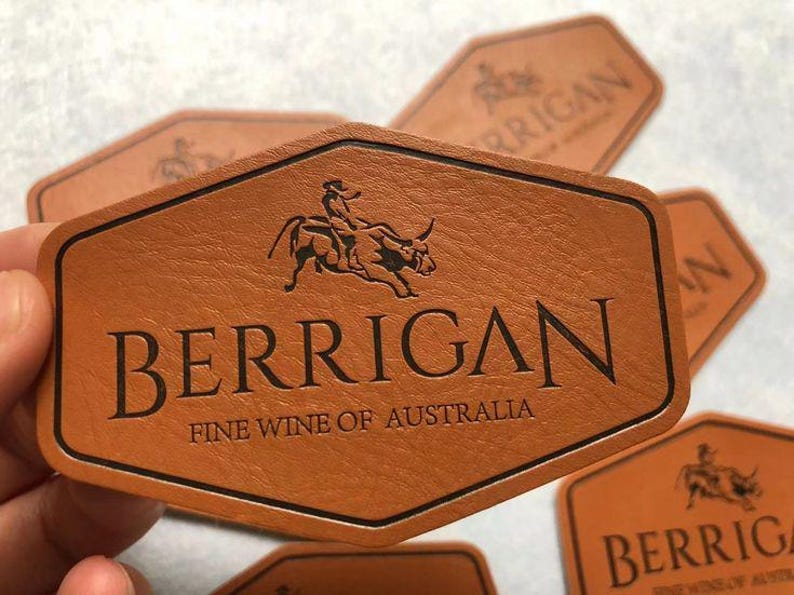 Może przedstawiać: Brązowa sk&oacute;rzana etykieta na wino z napisem "BERRIGAN" i tekstem "FINE WINE OF AUSTRALIA". Etykieta ma czarną obw&oacute;dkę i grafikę przedstawiającą osobę jadącą na byku. Etykieta ma sześciokątny kształt.