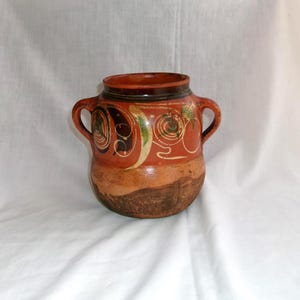 May include: A handcrafted, terracotta-colored ceramic pot with two handles. The pot features hand-painted designs in green, brown, and white. The base is a darker brown, and the pot has a wide opening. This rustic piece is ideal for home decor.