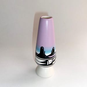 Peut inclure: Un petit vase en céramique de couleur mauve clair avec une base blanche. Le vase est décoré d'une scène de paysage désertique avec des montagnes et des cactus en noir et blanc.