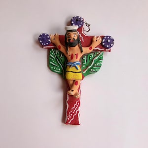 May include: A handcrafted clay crucifix featuring a figure with a beard, wearing a yellow loincloth with a blue bow. The cross is red with white accents, and the figure is attached to a green leaf-shaped base.
