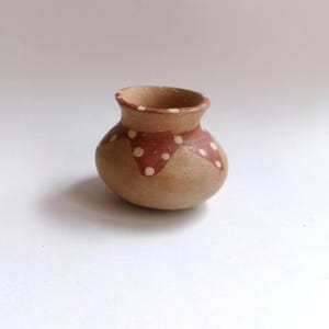 May include: A small, round, earthenware pot with a wide opening. The pot is tan with a band of dark red around the top, decorated with white polka dots. The pot is likely handmade and has a rustic appearance.