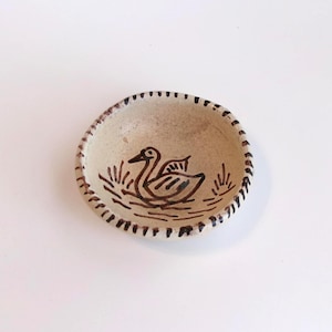 May include: A small, round, off-white ceramic bowl featuring a hand-painted dark brown illustration of a duck swimming in water. The rim of the bowl is decorated with a series of short, vertical dark brown lines encircling the entire edge.