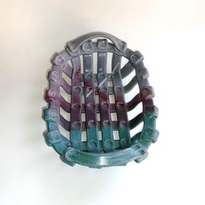 May include: A ceramic basket with a woven design in shades of purple, teal, and gray. The basket has a handle and is decorated with small, raised circles.