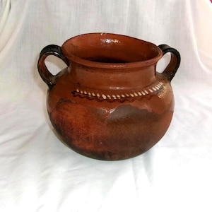 May include: A handcrafted, brown clay pot with two black handles. The pot features a decorative white dotted pattern around the rim. The rustic vessel is likely used for cooking or serving.