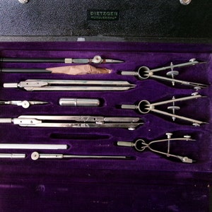 May include: A vintage Dietzgen Universal drafting compass set in a black leather case with a purple velvet interior. The set includes a variety of compasses, rulers, and other drafting tools.