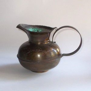 May include: A vintage copper pitcher with a round body and a wide, flared spout. The pitcher has a green patina on the inside and a worn, aged finish.