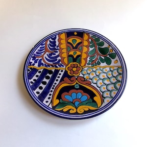May include: A round, hand-painted ceramic plate with a vibrant, multi-colored design. The plate features a blue border and is divided into sections with floral and geometric patterns in yellow, green, and blue. A central flower is also visible.