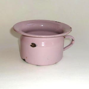 May include: A vintage, light pink chamber pot with a handle. The pot has a wide, flared rim and shows signs of wear, including small rust spots. The enamel finish is chipped in places, adding to its antique charm.