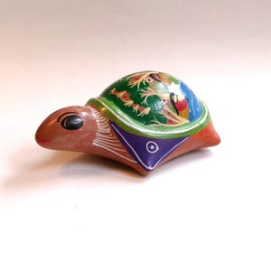May include: A small, hand-painted ceramic turtle figurine with a green, blue, and orange shell. The turtle has a brown body with black eyes and white details.