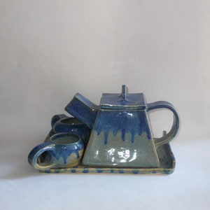 May include: A blue and green ceramic teapot with a lid and a matching tray. The teapot has a unique shape and is decorated with a drip glaze. There are three matching cups on the tray.