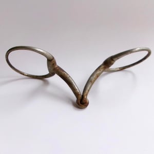 May include: An antique metal bit, featuring two oval rings connected by curved bars. The bit has a weathered, aged appearance with visible rust, suggesting it is an older equestrian item. The design is symmetrical and centered.