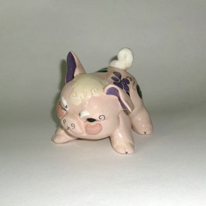 May include: A pink ceramic piggy bank with a curled tail, purple flowers, and green leaves painted on its side. The piggy bank has a winking face with black eyelashes and a pink nose.