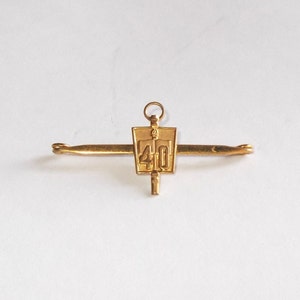 Puede incluir: Un broche de corbata dorado con un diseño rectangular que presenta el número "40" en el centro. El broche de corbata tiene un pasador en la parte posterior para sujetarlo a la ropa.