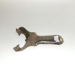 Puede incluir: Un abridor de botellas plateado vintage con un diseño curvo único. El abridor presenta un extremo con púas y una abertura circular, con el texto "EAST OAK LUMBER CO." visible. El metal muestra signos de edad y uso.