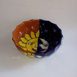 May include: A ceramic bowl with a sun and moon design. The sun is yellow with a smiling face and the moon is white with a crescent shape. The bowl is painted in shades of orange, yellow, blue, and white.