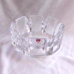 May include: A clear, faceted crystal bowl with a unique, geometric design. The bowl features a series of rectangular panels that create a flower-like shape. A small, red and white label is visible on the front. This decorative bowl is ideal for displaying small items.
