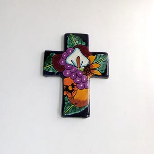 May include: A ceramic cross with a dark blue background, decorated with colorful floral and fruit motifs. The design includes a white calla lily, purple grapes, and a sunflower. The cross is a religious symbol.