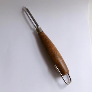 Puede incluir: Un pelador de verduras de acero inoxidable con mango de madera. El pelador tiene una cuchilla curva y un lazo en el extremo del mango. El mango es de madera marrón oscuro.