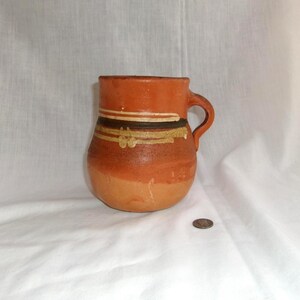 May include: A handmade, terracotta-colored mug with a handle. The mug features horizontal bands of brown, white, and gold paint around the middle. The bottom half of the mug is a lighter shade of terracotta. A coin is next to the mug.