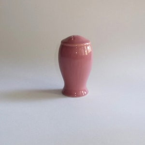 May include: A pink ceramic salt shaker with a rounded body and a small, slightly domed lid. The shaker has a classic, elegant shape and a glossy finish. The top of the lid has several small holes for dispensing salt.