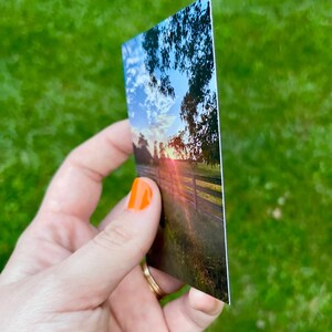 May include: A rectangular print depicting a sunset over a wooden fence and a field. The sky is a mix of blue, white, and orange hues. The print is held against a backdrop of green grass.