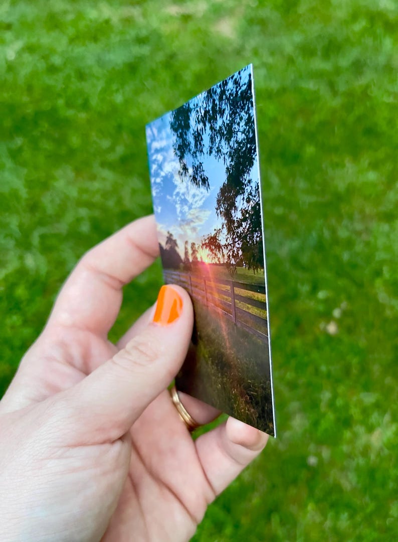 May include: A rectangular print depicting a sunset over a wooden fence and a field. The sky is a mix of blue, white, and orange hues. The print is held against a backdrop of green grass.