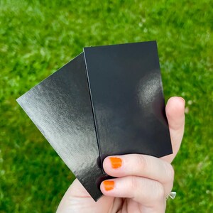 May include: Two black rectangular cards are held in a hand with orange nail polish. One card has a textured, metallic finish, while the other is smooth and glossy. The background is blurred green grass.