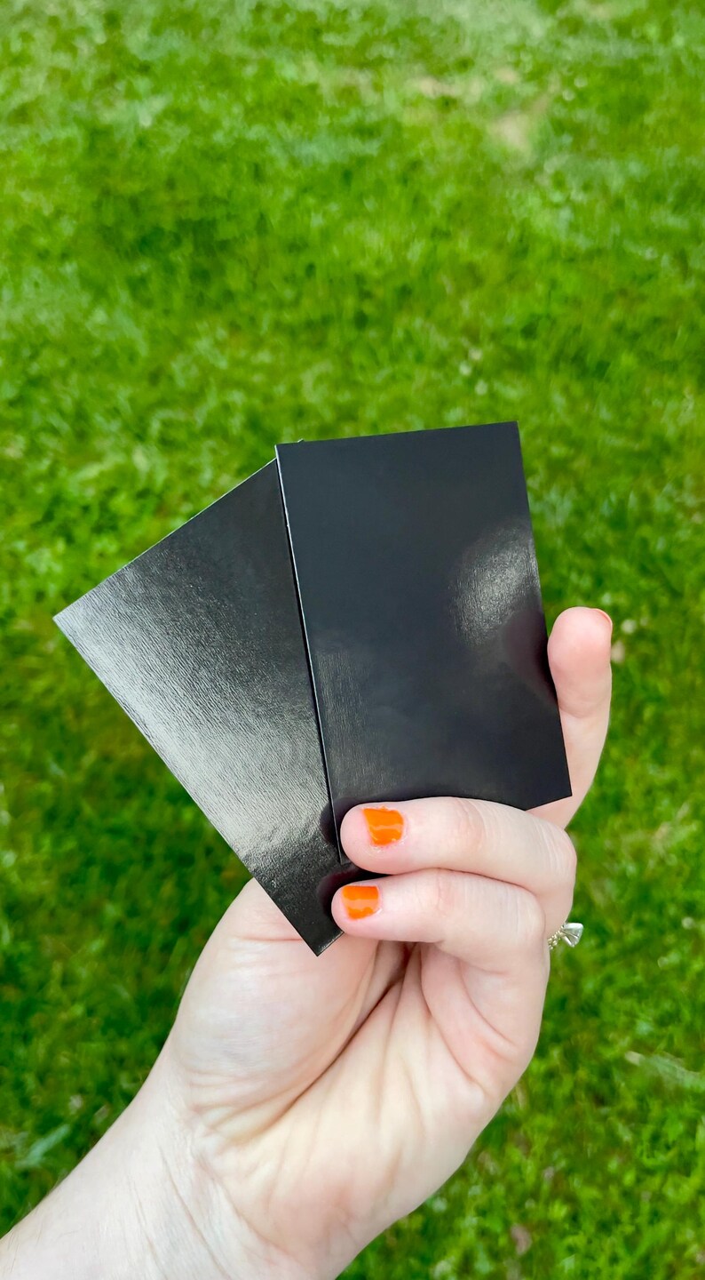 May include: Two black rectangular cards are held in a hand with orange nail polish. One card has a textured, metallic finish, while the other is smooth and glossy. The background is blurred green grass.