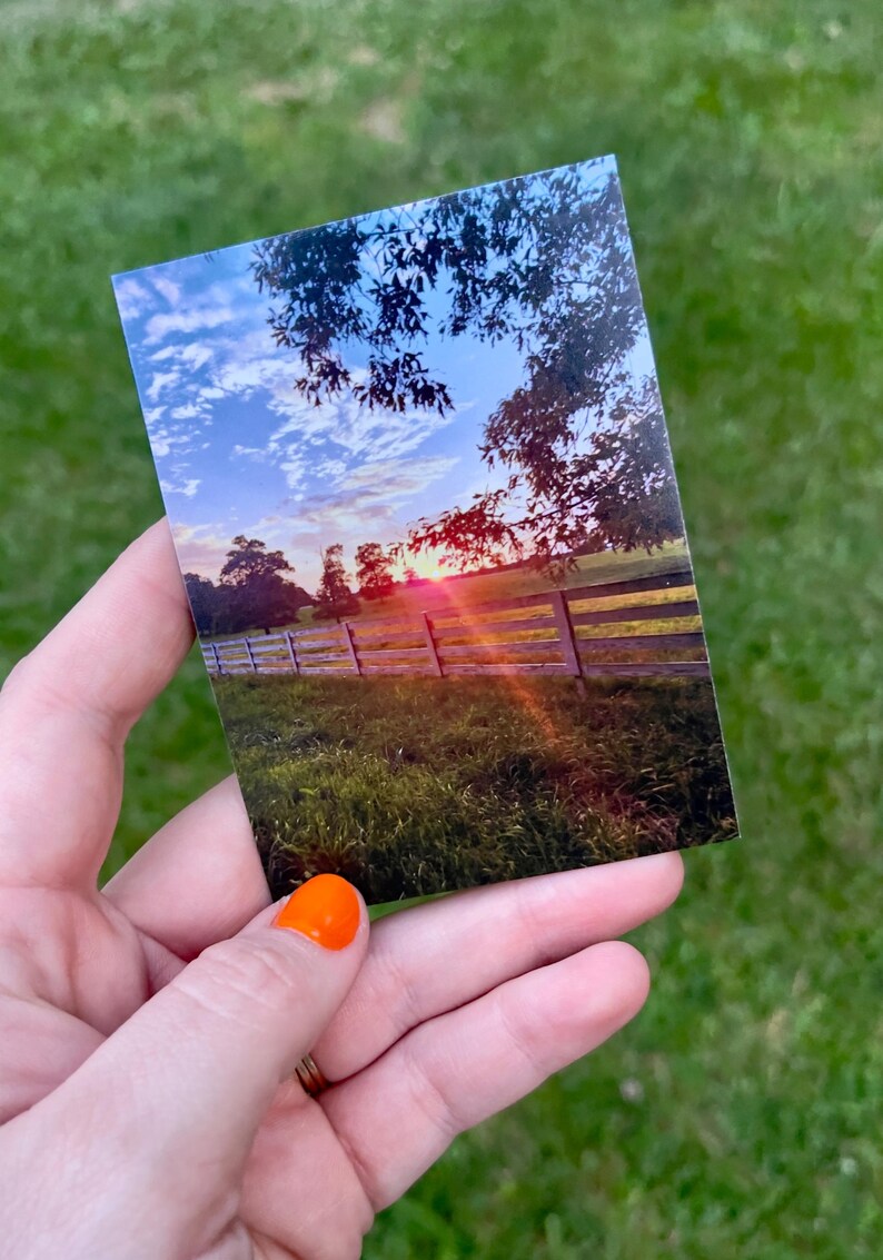 May include: A photograph of a sunset over a wooden fence in a field. The sky is a mix of blue, white, and orange hues. The sun is setting behind trees, casting a warm glow over the scene.