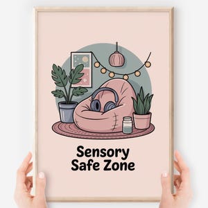 May include: A framed print with a light pink background features a cartoon illustration of a sensory safe zone. The image includes a pink beanbag chair, headphones, plants, and the text "Sensory Safe Zone."