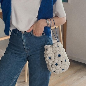 Könnte beinhalten: Eine weiße Häkeltasche mit blauen Blumenakzenten wird von einer Person getragen, die ein weißes T-Shirt und eine blaue Jeans trägt. Ein blauer Pullover ist über die Schultern gelegt. Die Tasche hat einen blauen Kordelzugverschluss.