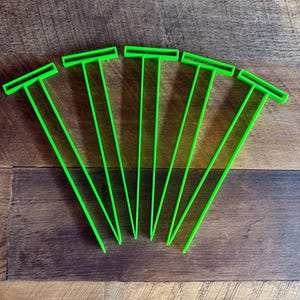 May include: Five bright green plant markers arranged on a wooden surface. Each marker has a T-shaped top and a long, thin stem. The markers are evenly spaced, creating a visually appealing pattern. These markers are ideal for organizing a garden.
