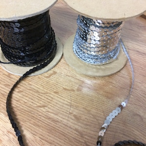 May include: Two spools of sequined trim, one black and one silver, on a wooden surface. The trim is made of small, round sequins.