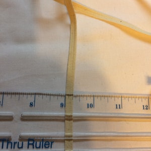 May include: A close-up of a beige cotton twill tape measuring 9 inches long. The tape is lying on a clear plastic ruler with the words "Thru Ruler" printed on it.