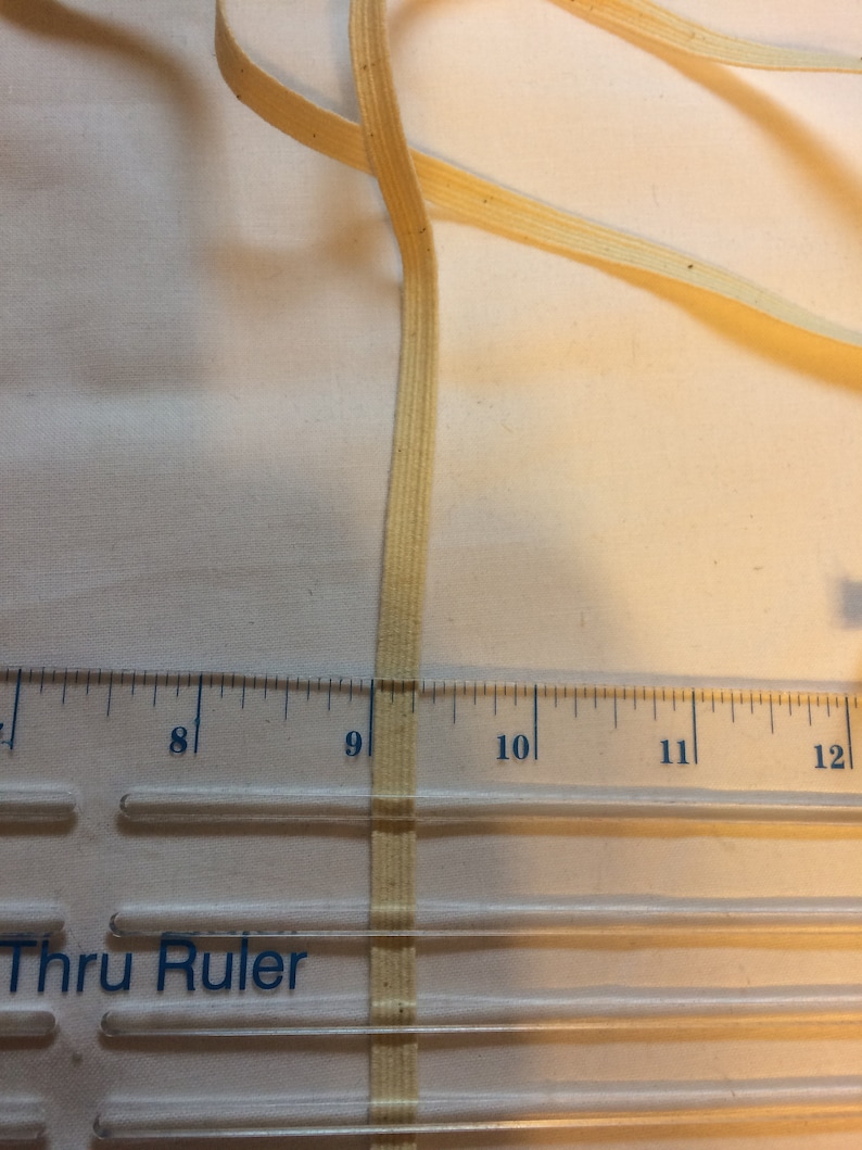 May include: A close-up of a beige cotton twill tape measuring 9 inches long. The tape is lying on a clear plastic ruler with the words "Thru Ruler" printed on it.