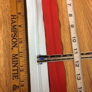 May include: Two zipper tapes, one white and one red, are laid out next to each other. A ruler is placed on top of the zipper tapes to show the length. The ruler is marked in inches and centimeters.