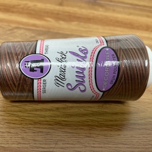 May include: A spool of brown and white thread with the text "Maxi-Lock Serger Thread Swirls Decorative Spun Polyester" on the label.
