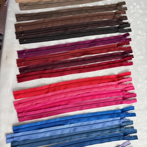 May include: A collection of long nylon zippers arranged in horizontal rows, featuring a gradient of colors including beige, brown, dark brown, burgundy, red, pink, blue, and purple, each with a metal pull tab at the top end of the tape.