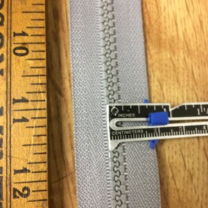 May include: A close-up of a silver zipper tape with a zipper slider. A ruler is used to measure the zipper tape, which is 1 inch wide.