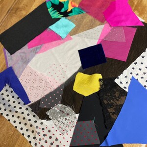 May include: A selection of fabric swatches in diverse colours and textures. Includes black, pink, blue, white, and patterned fabrics. Some swatches feature sequins, polka dots, or floral prints. The fabrics are arranged on a wooden surface.