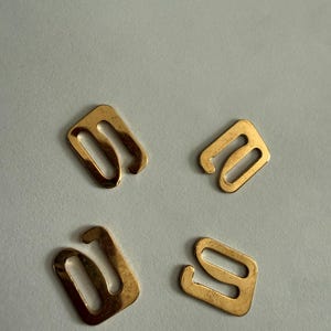 May include: Four small, gold-colored metal bra strap hooks are arranged on a light gray surface. Each hook has a rectangular base with a curved, open end, designed to attach to a bra strap.