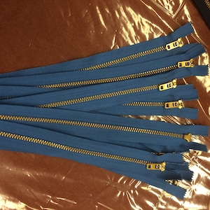 May include: A close-up of a bundle of blue zipper tape with gold metal teeth. The zippers are arranged in a stack, with the teeth facing up.