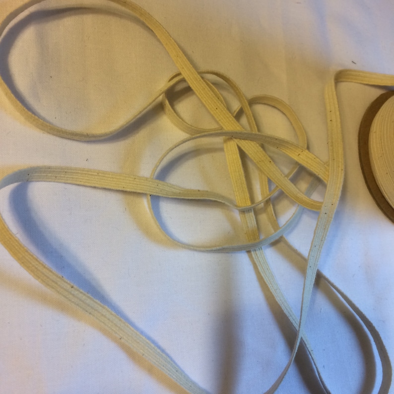 May include: A close-up of a beige cotton elastic cord. The cord is folded and twisted, showing its texture and flexibility.
