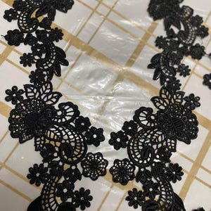 May include: Two black lace appliques with floral designs. The appliques are delicate and intricate, with a variety of flower shapes and sizes.